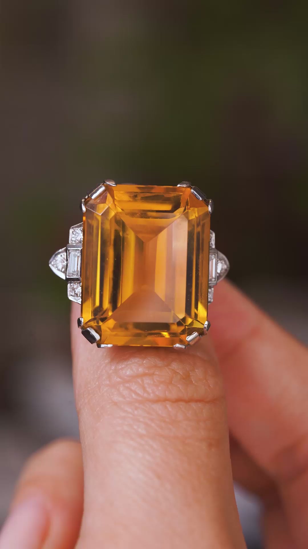 A video of a platinum Antique, Art Deco citrine and diamond cocktail ring held in between fingers and slowly rotated to show off different angles against a blurred background