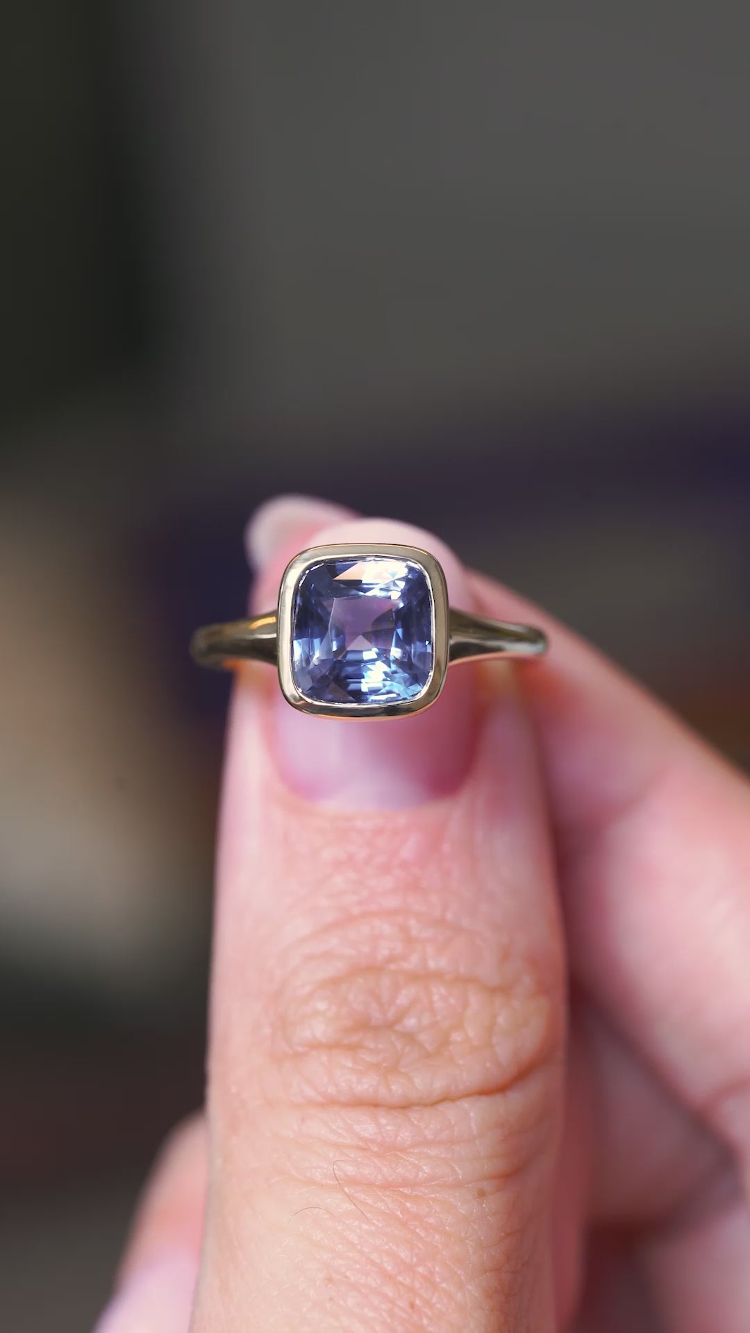 A video of a yellow gold antique-inspired blue sapphire solitaire ring held in between the fingers and turned to showcase different angles against a blurry background.