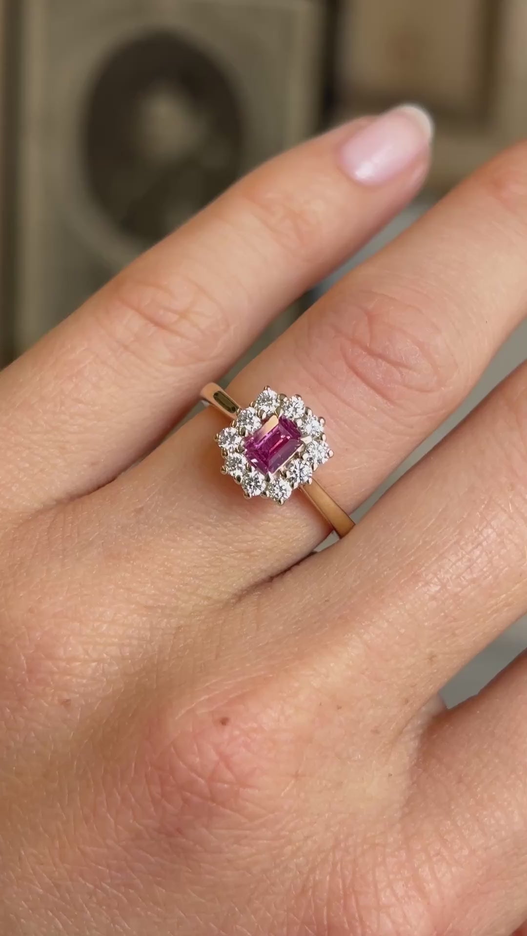 Antique-inspired yellow gold ruby and diamond cluster ring worn on hand, featuring a vibrant step-cut pinkish-red ruby surrounded by brilliant diamonds, filmed against a softly lit vintage interior backdrop.