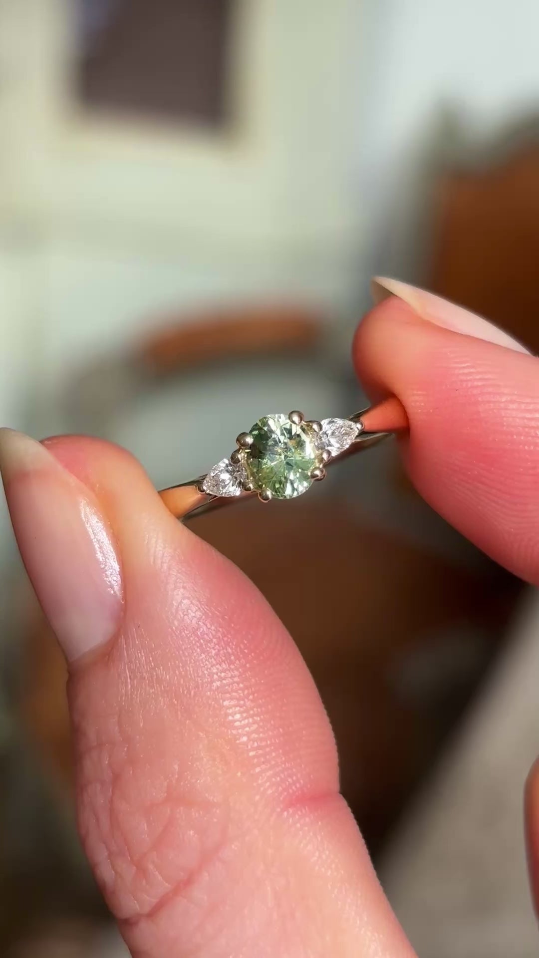 An engagement ring showing an small oval-cut greeny-teal sapphire and diamond three-stone engagement ring, captured between two fingers being shown in natural daylight