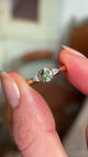 An engagement ring showing an small oval-cut greeny-teal sapphire and diamond three-stone engagement ring, captured between two fingers being shown in natural daylight