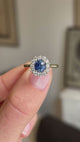 A close-up video of an antique-inspired, blue sapphire at the centre with a diamond halo cluster set in 14ct yellow gold held between fingers.