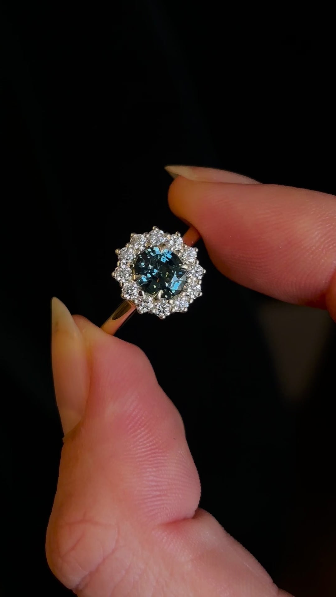 Teal sapphire and diamond cluster yellow gold engagement ring in fingers, shown in motion to indicate scale.