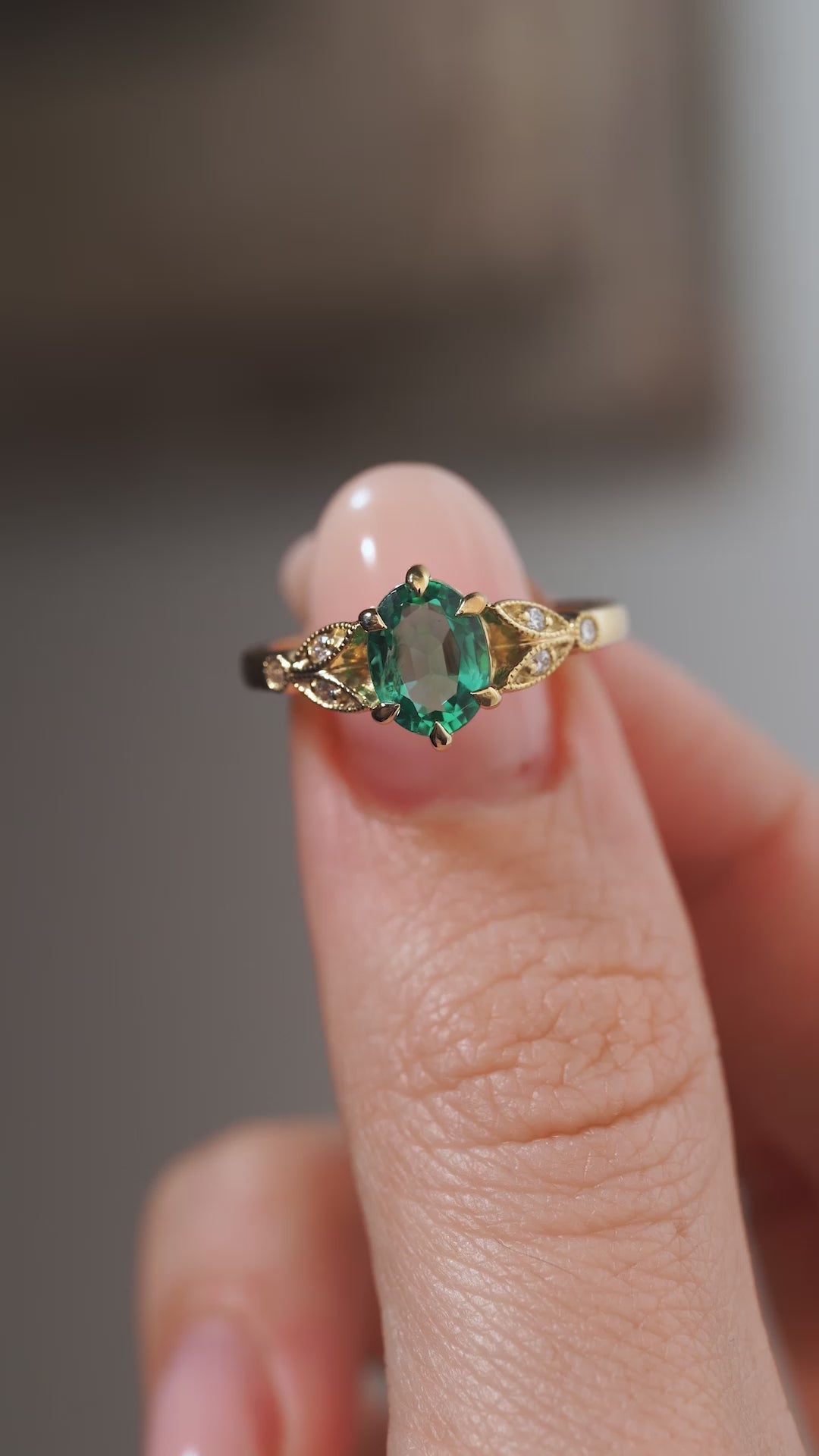 A close-up video of a vintage-inspired emerald and diamond engagement ring held between fingers, showcasing the rich green gemstone, delicate diamond accents and intricate yellow gold detailing.