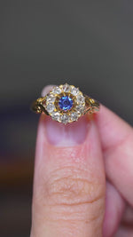 A video of an Antique, Victorian sapphire and old mine-cut diamond cluster engagement yellow gold ring held between the finger and the thumb and turned to showcase different angles against a blurry grey background.