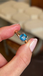 A video of an Antique-inspired, teal square-cut sapphire solitaire engagement ring in yellow gold held between two fingers and rotated slowly to display all profiles of the ring
