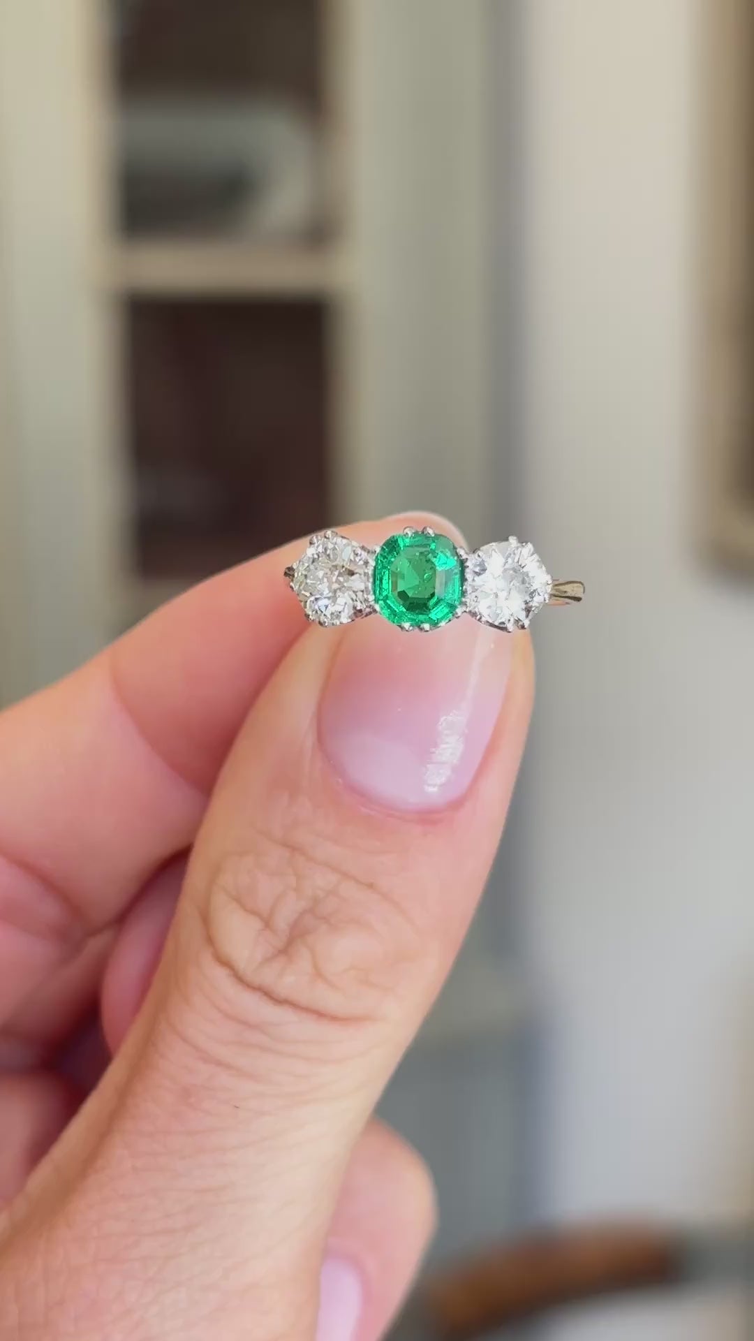 Antique Edwardian emerald and diamond three-stone engagement ring showcasing an emerald centre stone and diamond side stones, held by hand to highlight the setting and craftsmanship.