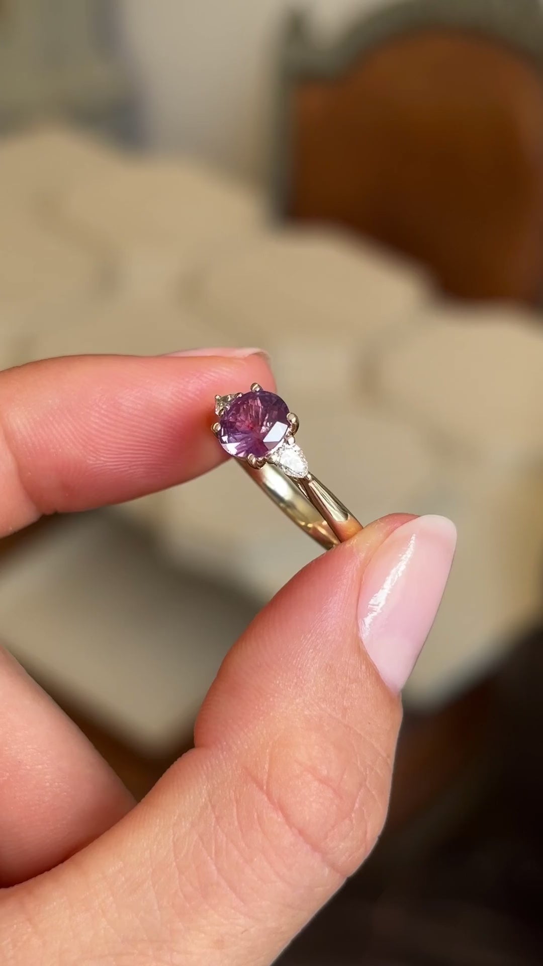 Pink-red sapphire and diamond three-stone engagement ring featuring a round pink-red sapphire centre stone and pear-cut diamond accents, held by hand for detailed viewing.
