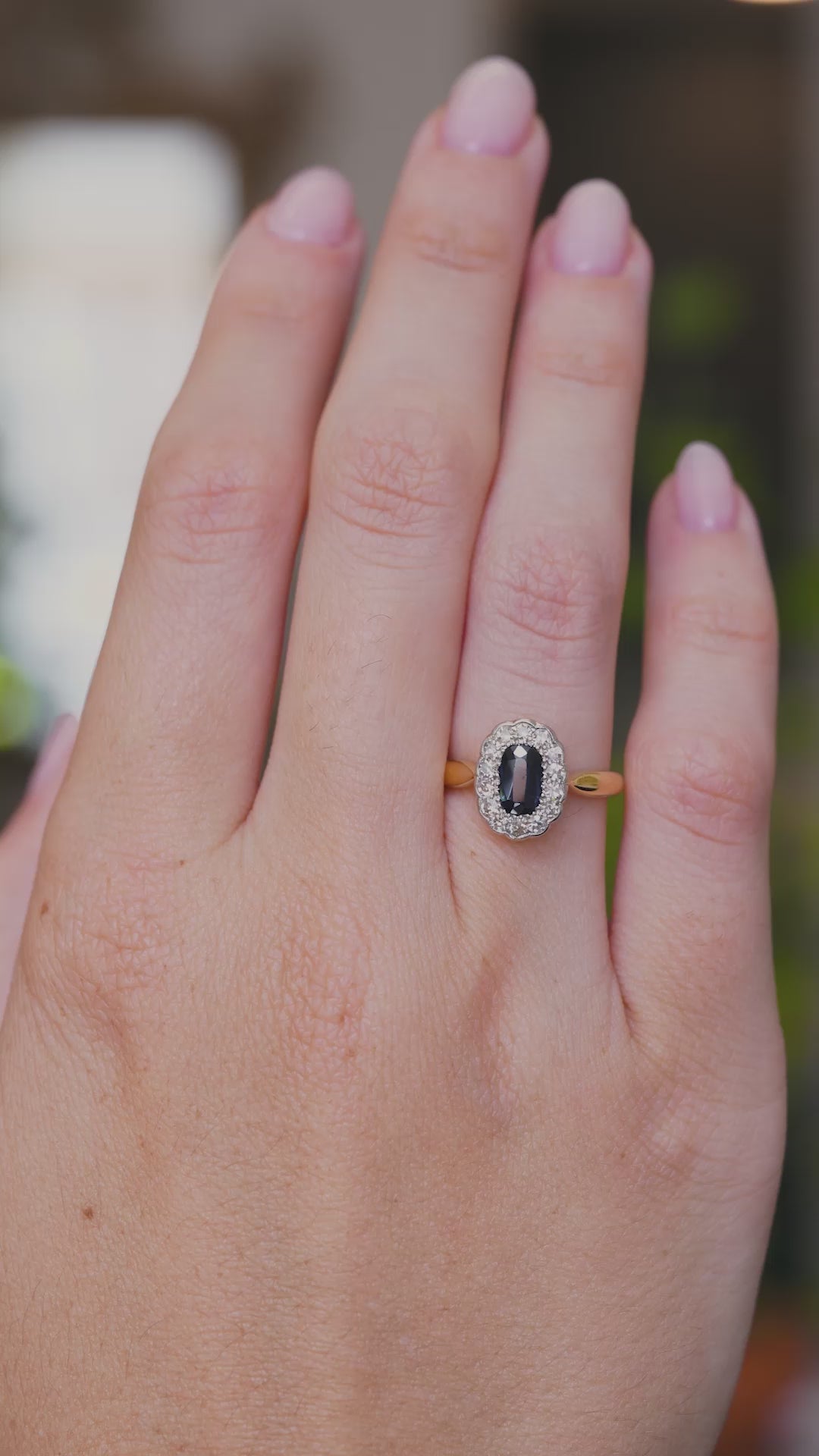 A video of a yellow gold Antique, Edwardian sapphire and diamond coronet cluster engagement ring worn on the hand  as the fingers move, shown against a blurred background.