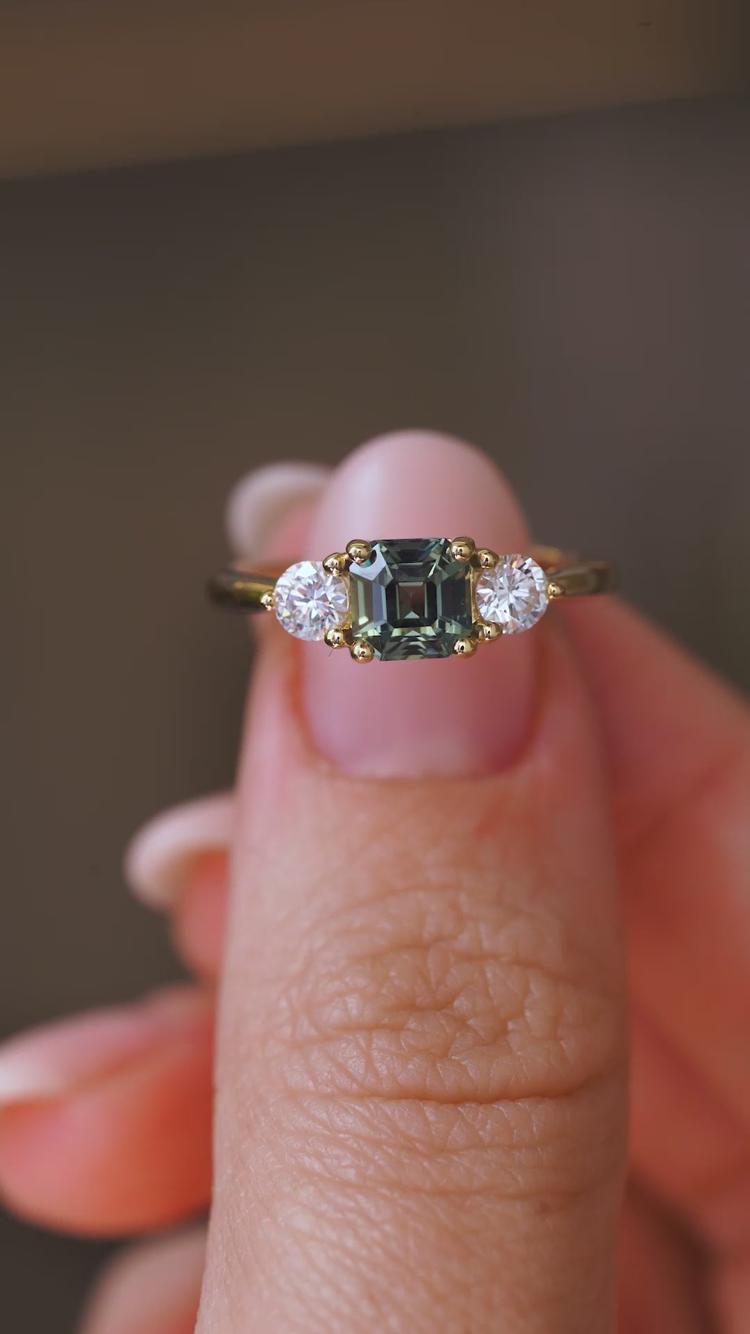 A video of an Antique-inspired, Teal sapphire and diamond three-stone yellow gold engagement ring held in between the fingers and rotated to show angles
