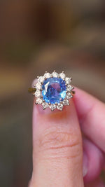 A vintage-inspired Ceylon intense blue sapphire and diamond halo engagement ring shown in video between fingers. 