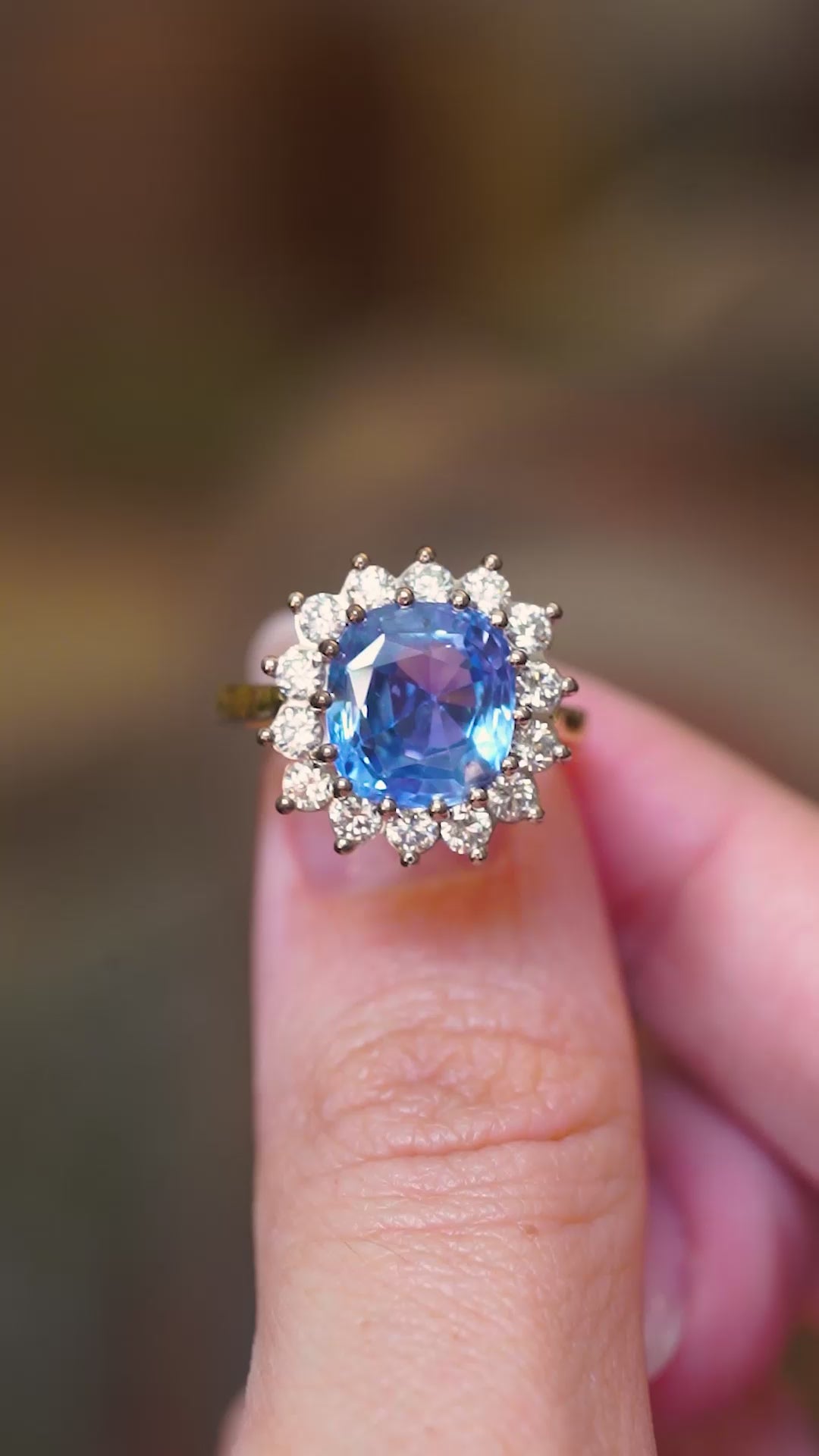A vintage-inspired Ceylon intense blue sapphire and diamond halo engagement ring shown in video between fingers. 
