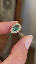 Antique-inspired pear-cut teal tourmaline and diamond cluster engagement ring with a pear-shaped teal tourmaline centre stone and sparkling diamond halo, held by hand for detailed viewing.