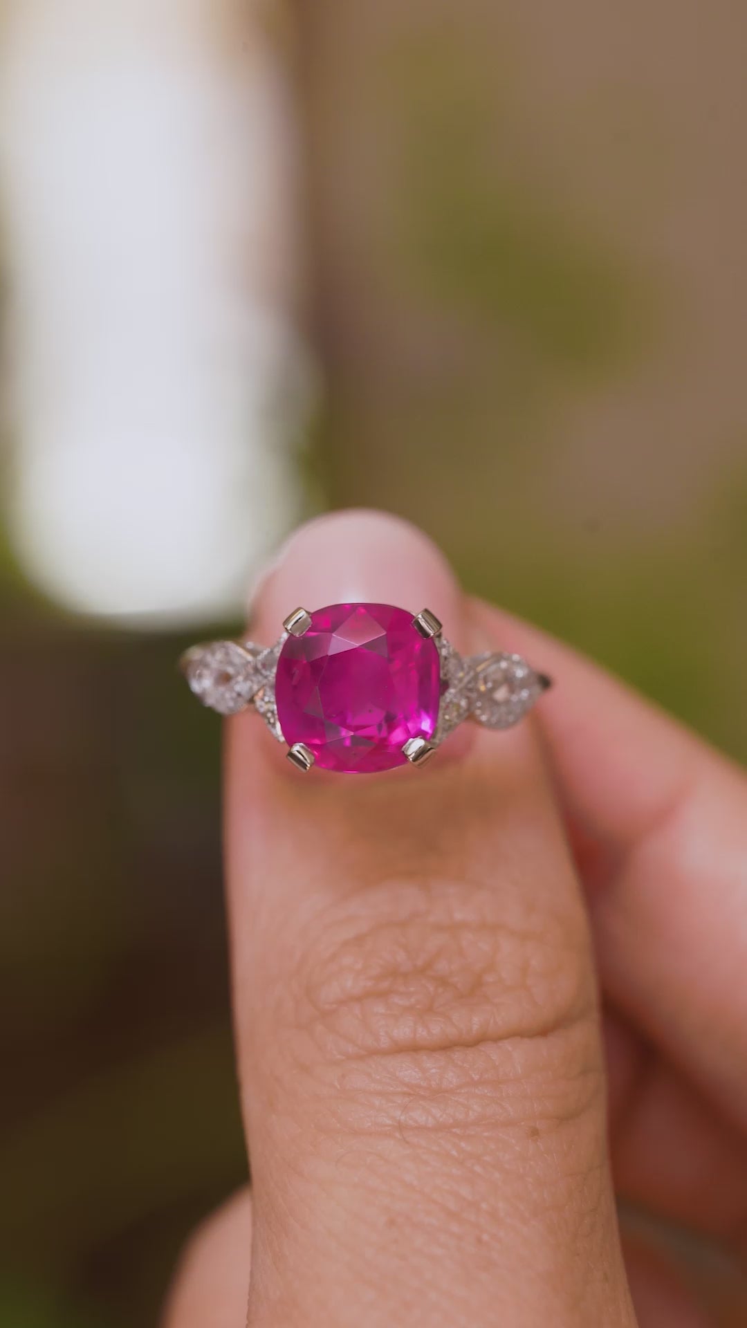 A close up video of an Antique white gold 1920s Art Deco ruby flanked solitary engagement ring held between two fingers and rotated to see all the profiles of the ring and the setting of the central Burmese pink ruby.