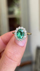 Detailed video of an oval-cut emerald and diamond cluster ring in yellow gold, held between fingers to showcase the vivid green emerald, diamond halo and elegant antique-inspired design against a warm interior setting.