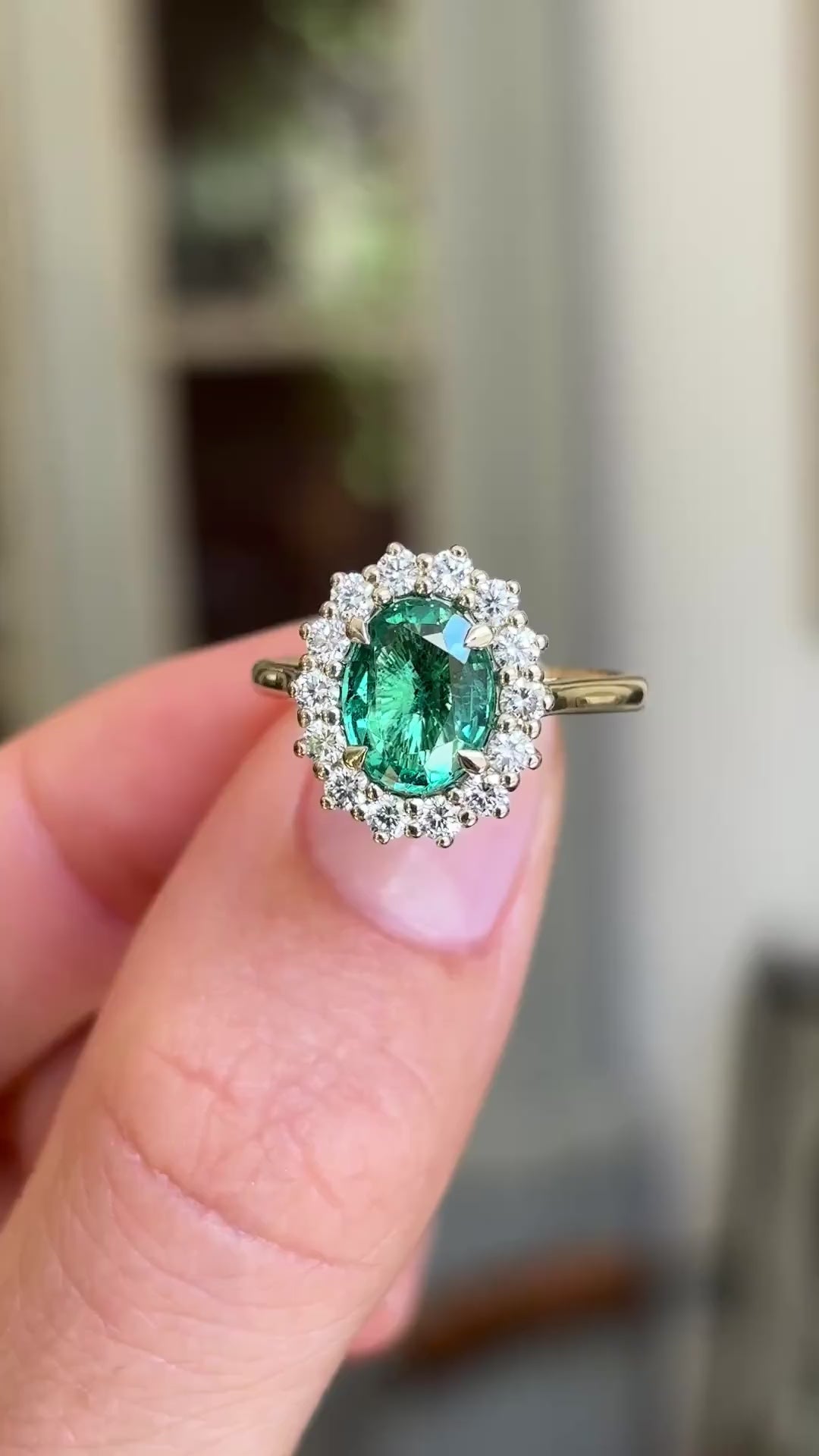 Detailed video of an oval-cut emerald and diamond cluster ring in yellow gold, held between fingers to showcase the vivid green emerald, diamond halo and elegant antique-inspired design against a warm interior setting.