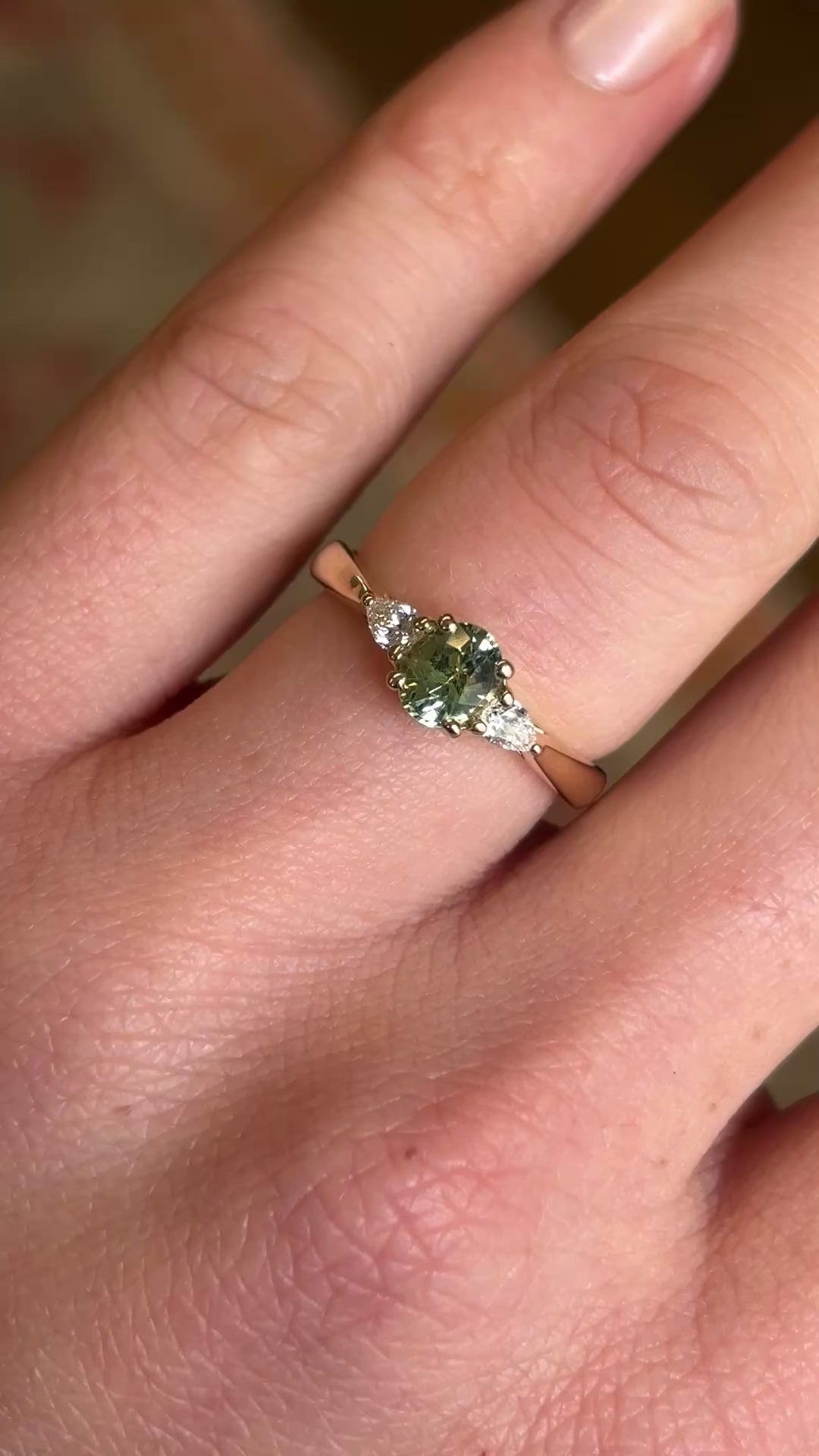 Close-up of a three-stone engagement ring with an oval-cut greeny-teal sapphire and diamond accents, worn on the hand
