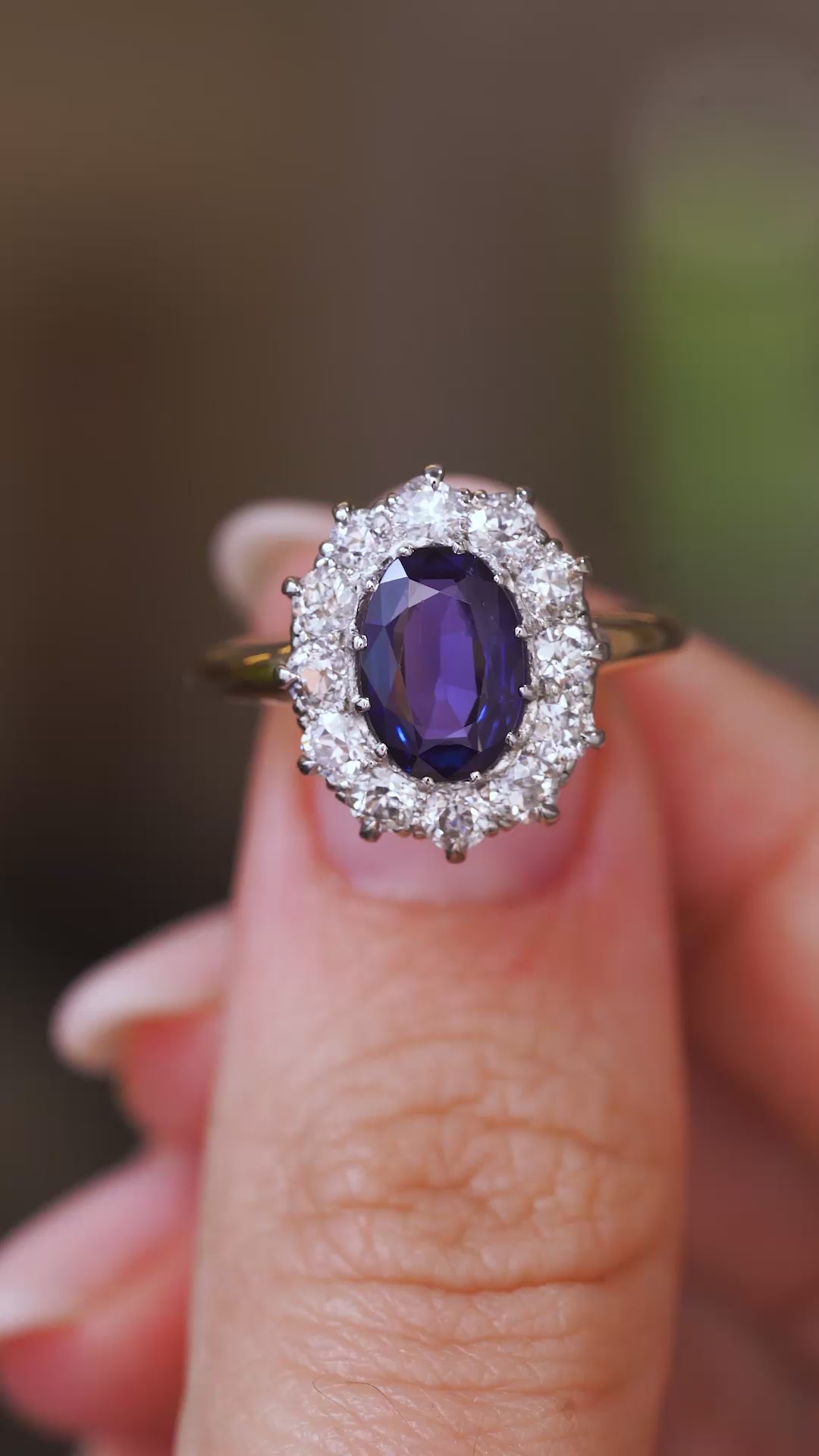 A close up video of a Vintage, 1940s blue sapphire and diamond cluster engagement ring held between two fingers and rotated slowly to show the front and side profiles of the ring and the yellow gold band