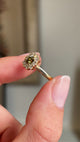 Close up video of the side profiles of an Antique-inspired, Brown diamond cluster engagement ring, showcasing the central square cut diamond and halo of diamonds surrounding it.  Held between two fingers against a blurred background. 