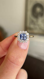 A close up video of an Antique-inspired, cluster engagement ring in yellow gold, held between two fingers and rotated slowly to showcase the central cushion-cut blue sapphire and surrounding cluster of diamonds. 