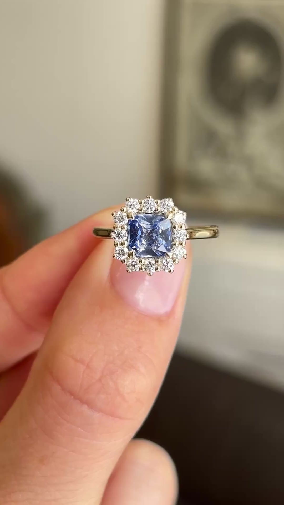 A close up video of an Antique-inspired, cluster engagement ring in yellow gold, held between two fingers and rotated slowly to showcase the central cushion-cut blue sapphire and surrounding cluster of diamonds. 