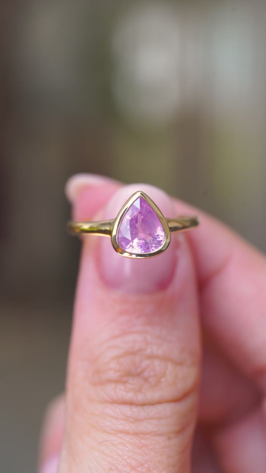 A video of an antique-inspired yellow gold engagement ring featuring a pear-cut pink sapphire bezel set single stone style held in the fingers and turned to show different angles.