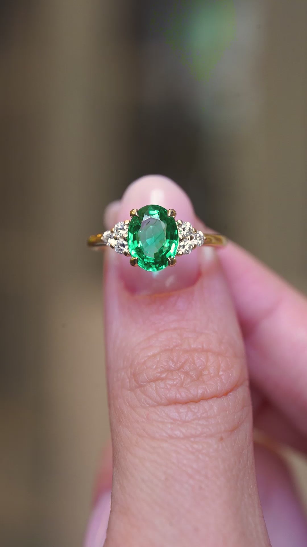 A video of vintage-inspired oval-cut emerald and diamond trefoil engagement ring held between the fingers.