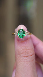 A video of vintage-inspired oval-cut emerald and diamond trefoil engagement ring held between the fingers.