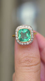 A video of an antique Belle Époque cushion-cut emerald and diamond cluster engagement ring in yellow gold held between the fingers and rotated to show different angles of the ring against a blurry background. 