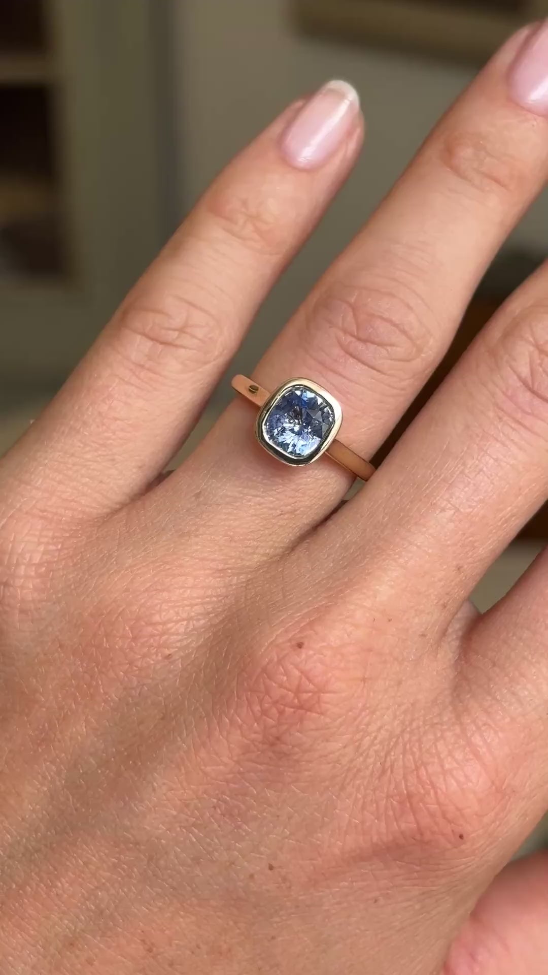 Video of an Antique-inspired, blue  cushion-cut sapphire solitaire engagement ring in yellow gold worn on a wedding finger against a blurred background. 
