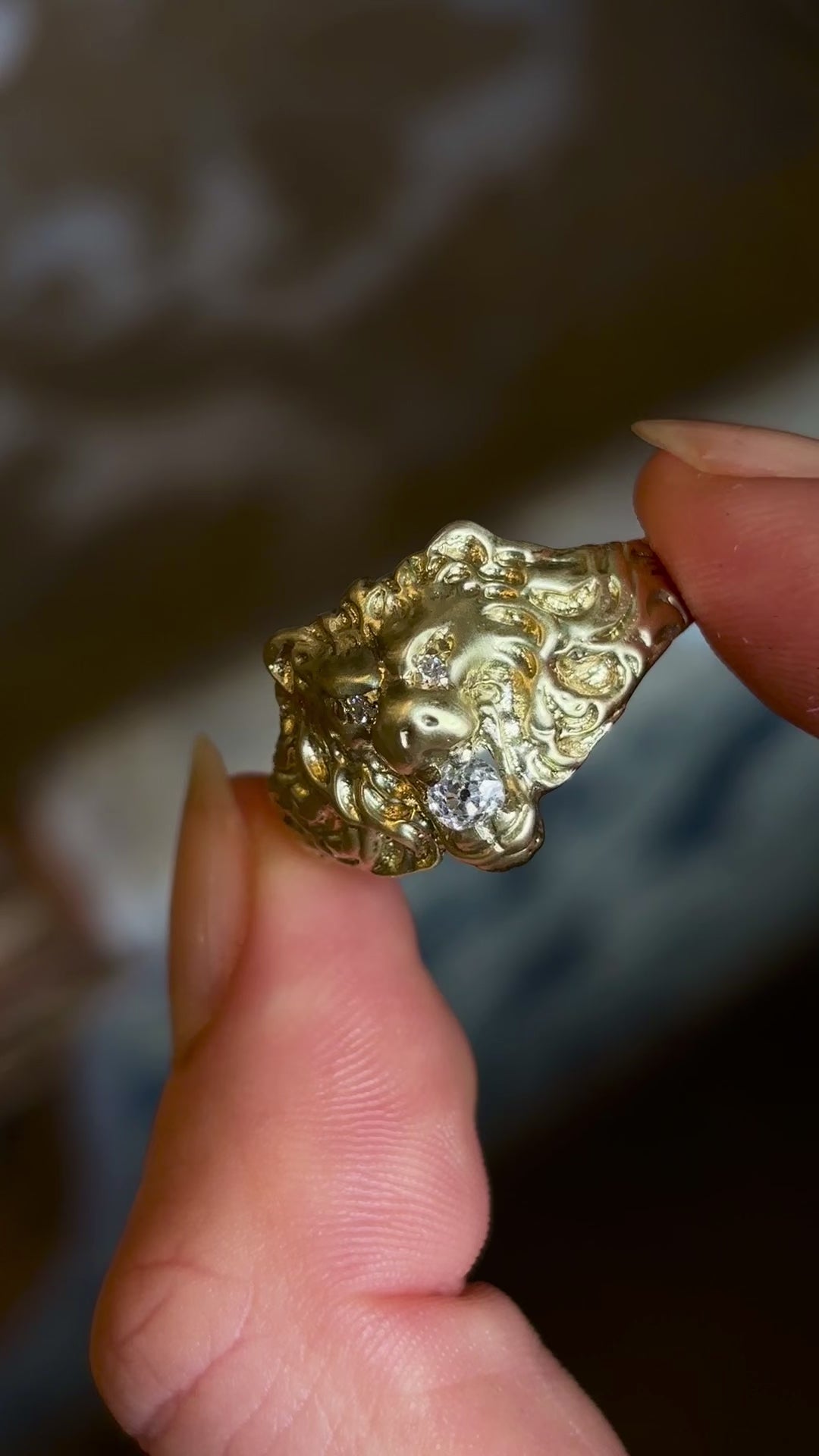 Vintage 1950s old-cut diamond lion’s head ring with diamond-set eyes held in fingers and rotated slowly, on a blurred background. 