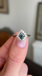 A close up video of an Antique-inspired, green sapphire and diamond cluster engagement ring in gold, held between two fingers and rotated to display all the side profiles of the ring
