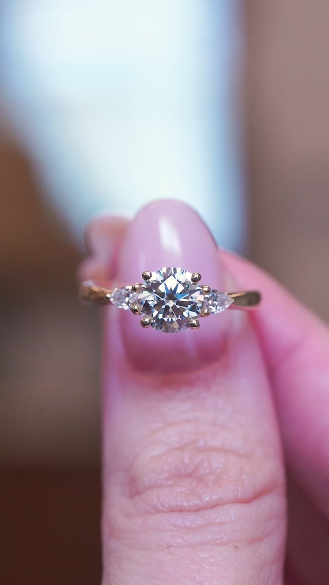 Round brilliant-cut diamond three-stone engagement ring held between fingers showing ring details.