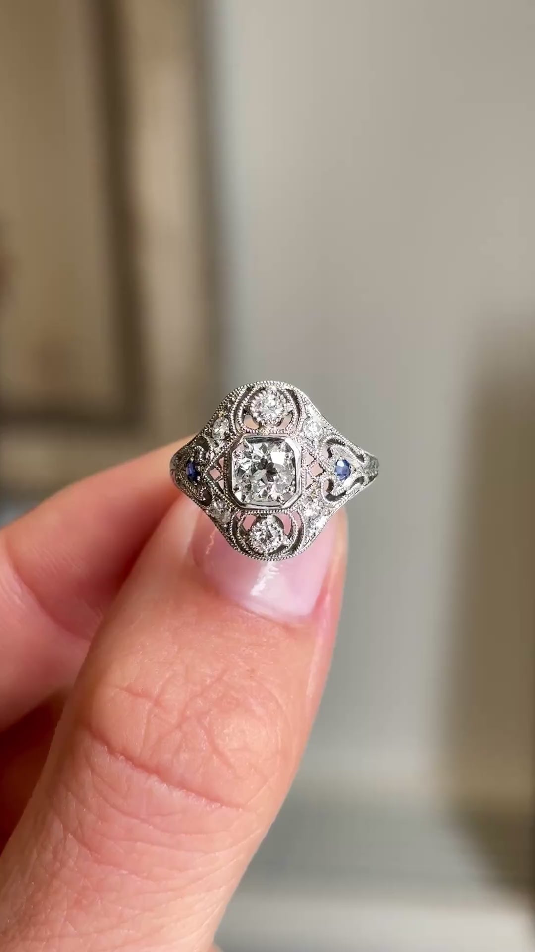 Detailed video of an antique 1920s platinum sapphire and diamond cluster ring, highlighting the rich blue sapphire centre stone, brilliant diamond surround and elegant Art Deco craftsmanship against a warm interior setting.