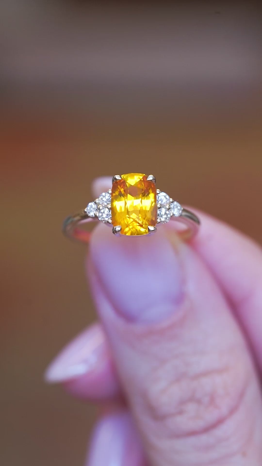 Orange cushion-cut sapphire and diamond cluster trefoil ring held between fingers showing gemstone sparkle.