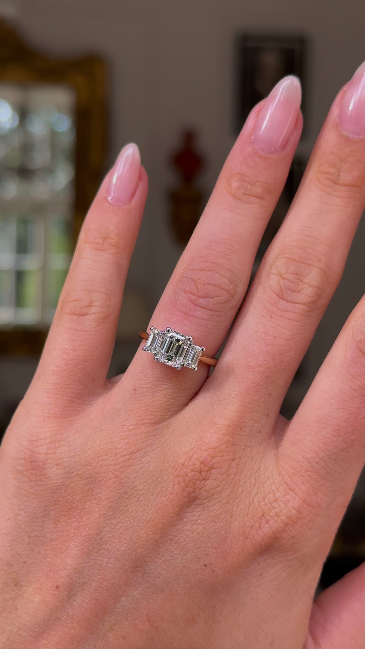 Three stone emerald-cut diamond ring, worn on left hand, ring finger. Hand moving away from lens, hot with antiquated bokeh background