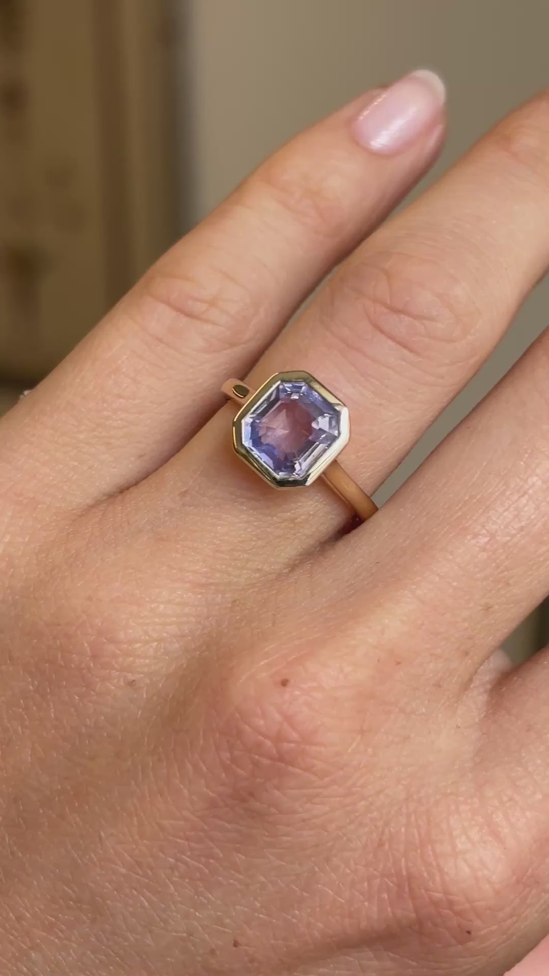 Antique-inspired yellow gold solitaire engagement ring worn on hand, featuring a square lavender sapphire in a smooth rub-over setting, filmed against a softly lit vintage interior backdrop.
