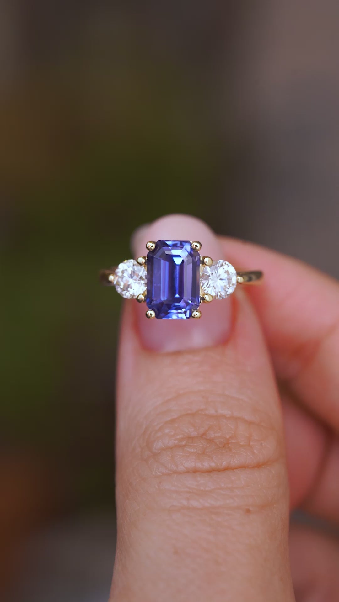 A close up video of an Antique-inspired, blue sapphire and diamond three-stone engagement ring in yellow gold.  Held between two fingers and rotated slowly to showcase the elongated cushion-cut of the blue sapphire and side profile of the ring.