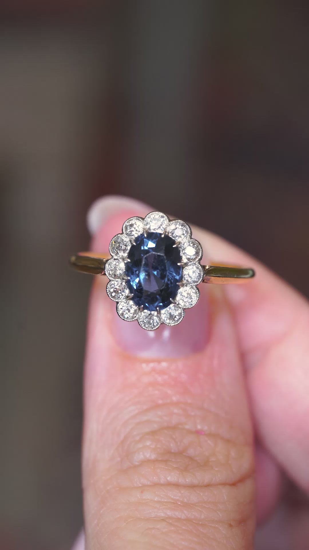 A video of an Antique, Edwardian blue spinel surrounded by a halo of diamonds in a coronet cluster engagement ring in yellow gold held in the hand and turned to showcase different angles. 