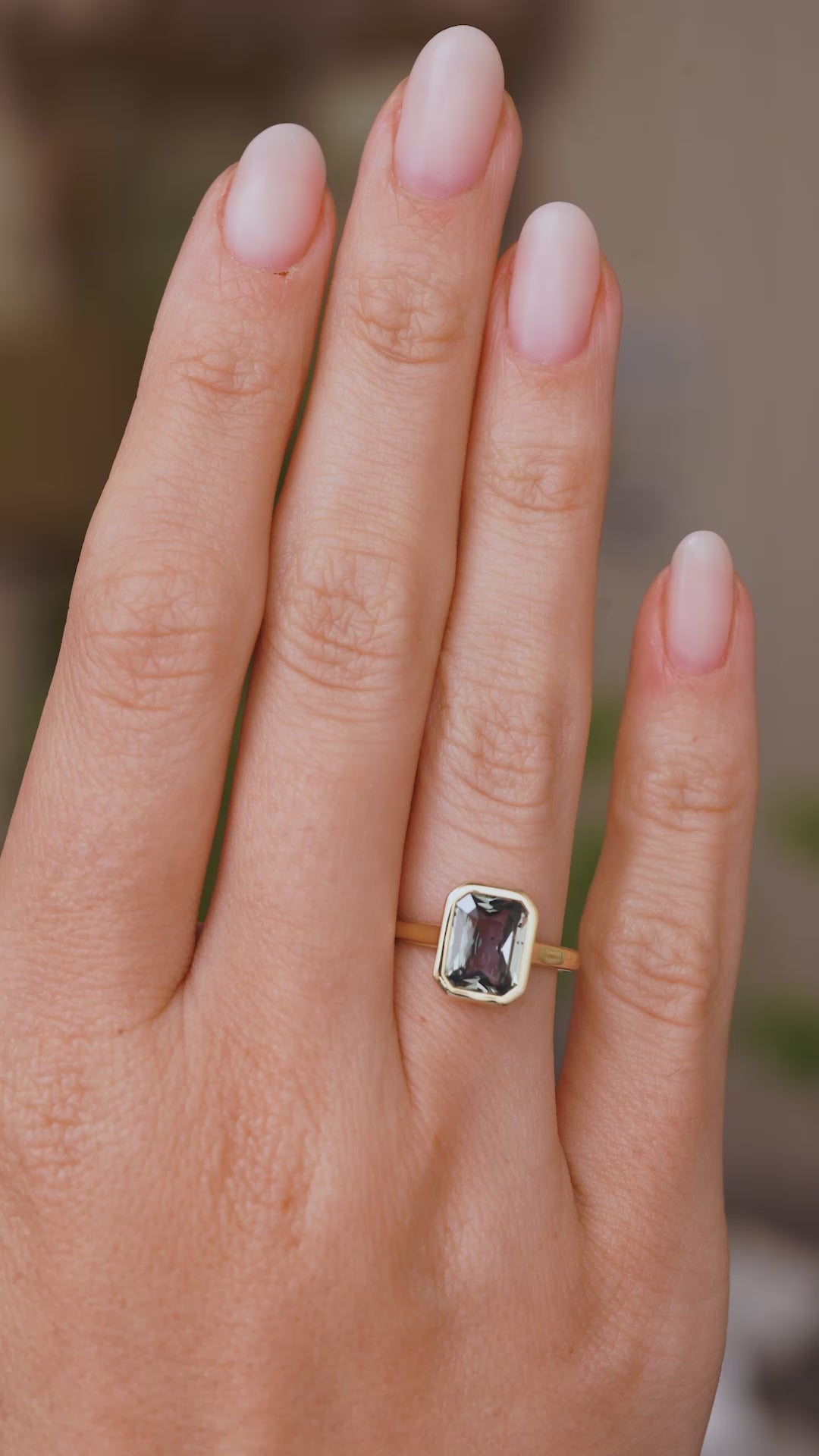 A close up video of a hand wearing a sapphire solitaire ring on a wedding finger.  Rotated slowly to showcase the Antique-inspired design, low profile of the yellow gold setting and bezel set blue-grey rectangular cut sapphire. 