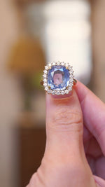 Close-up of an antique-inspired soft blue sapphire and diamond cluster ring in yellow gold held between fingers. 