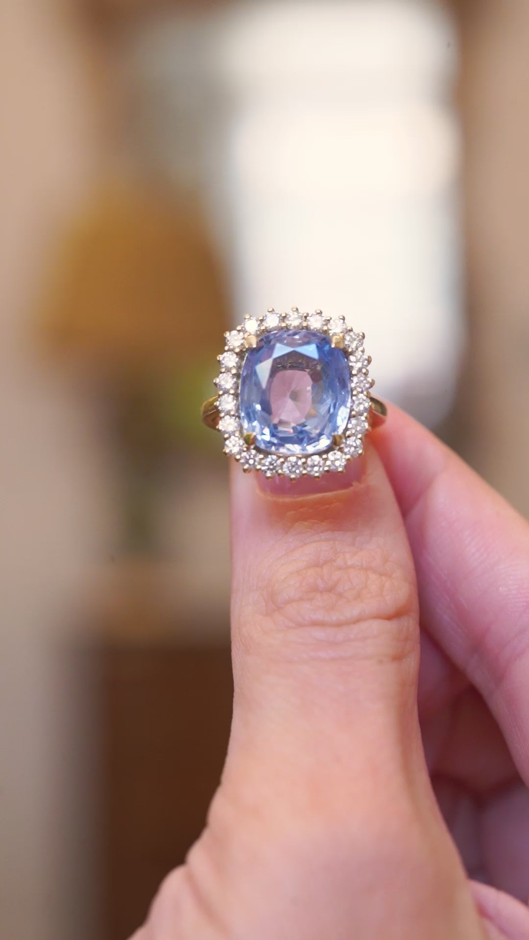 Close-up of an antique-inspired soft blue sapphire and diamond cluster ring in yellow gold held between fingers. 