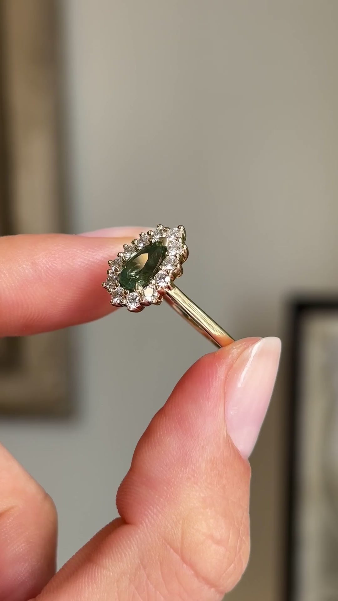 Detailed video of an antique-inspired green sapphire and diamond halo ring in yellow gold, held between fingers to showcase the pear-shaped centre stone, diamond cluster setting and vintage-inspired design.