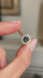 An Antique-inspired yellow gold engagement ring held between fingers, showcasing a teal sapphire and diamond cluster.