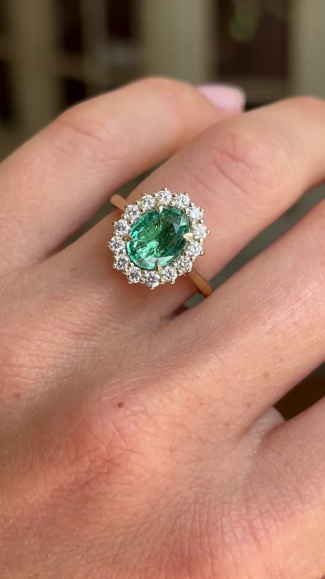 Antique-inspired yellow gold emerald and diamond cluster ring worn on hand, featuring a vibrant oval-cut emerald surrounded by brilliant diamonds, filmed against a softly lit vintage interior backdrop.