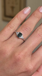 A video of a vintage-inspired engagement ring with a green sapphire at the centre, flanked by a diamond cluster trefoil on both shoulders. 