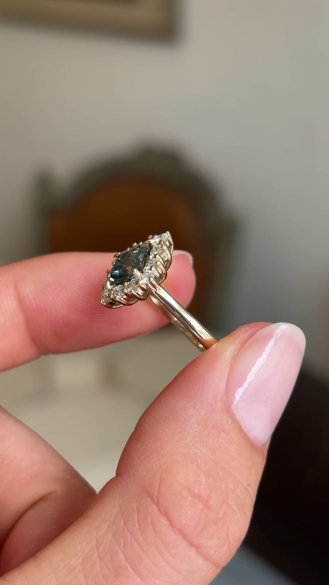 A close up video of an Antique-inspired, Teal kite-cut sapphire and diamond cluster engagement ring in gold held between two fingers against a blurred background
