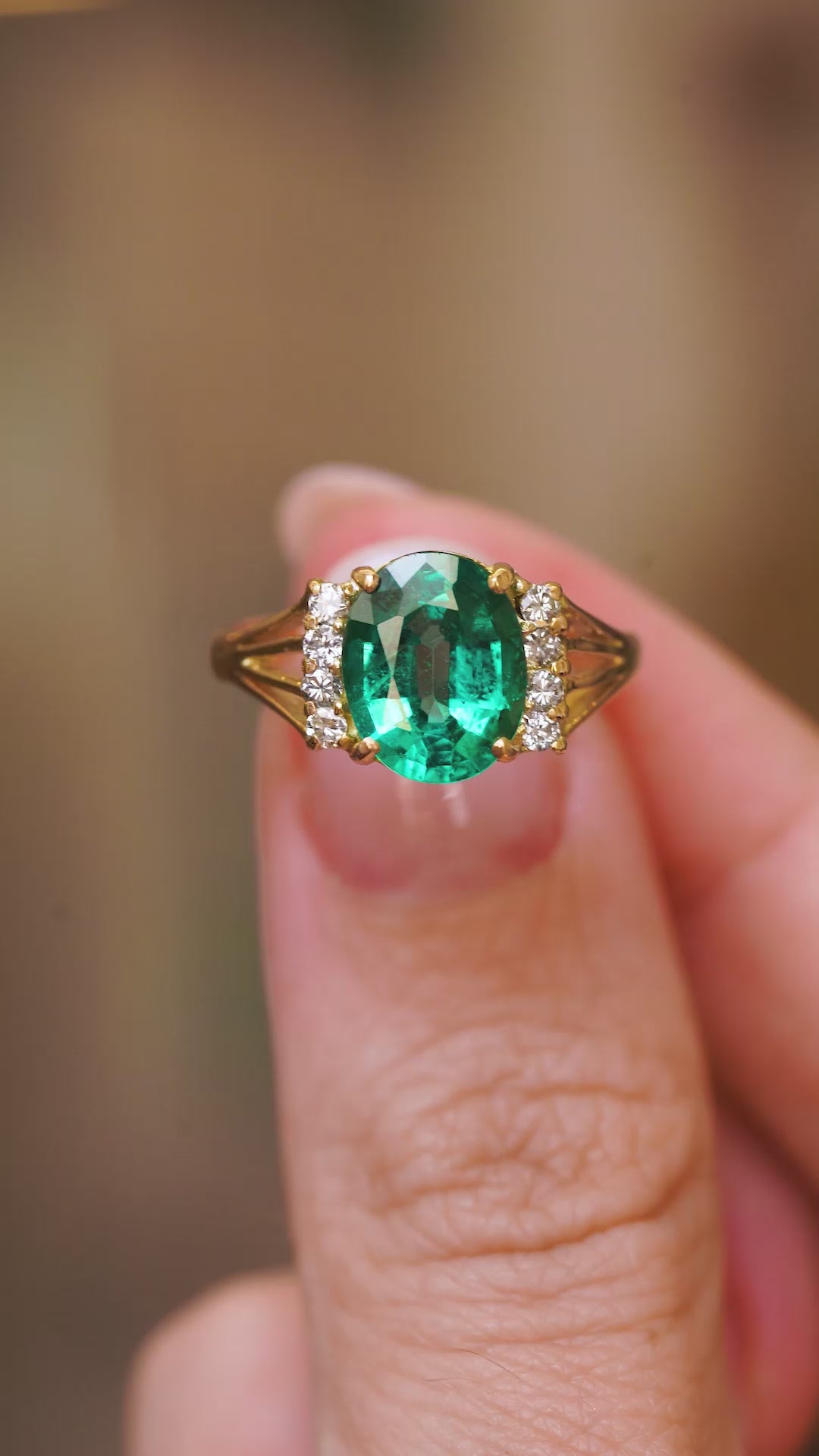 A close up video of a Vintage, 1970s emerald and diamond engagement ring in yellow gold, showcasing the large central green stone and diamonds that flank it on either side.  Held between two fingers and rotated to showcase the profiles of the ring. 