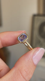 Detailed video of a square lavender sapphire solitaire ring in yellow gold, held between fingers to showcase the smooth rub-over setting, soft violet-blue tones and elegant antique-inspired design against a warm interior setting.