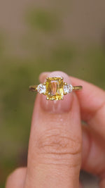 A video of a vintage style, Yellow sapphire and diamond three-stone engagement ring in yellow gold held between the fingers against a blurry background.